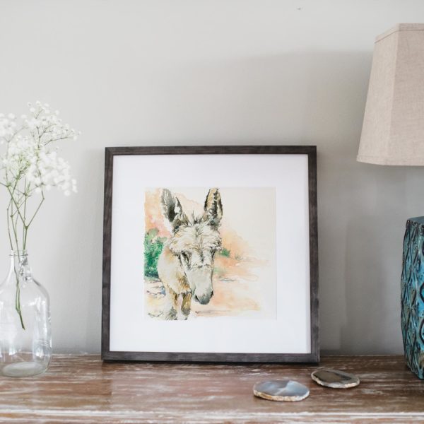 Watercolor of a donkey in Petra in a gray frame next to flowers